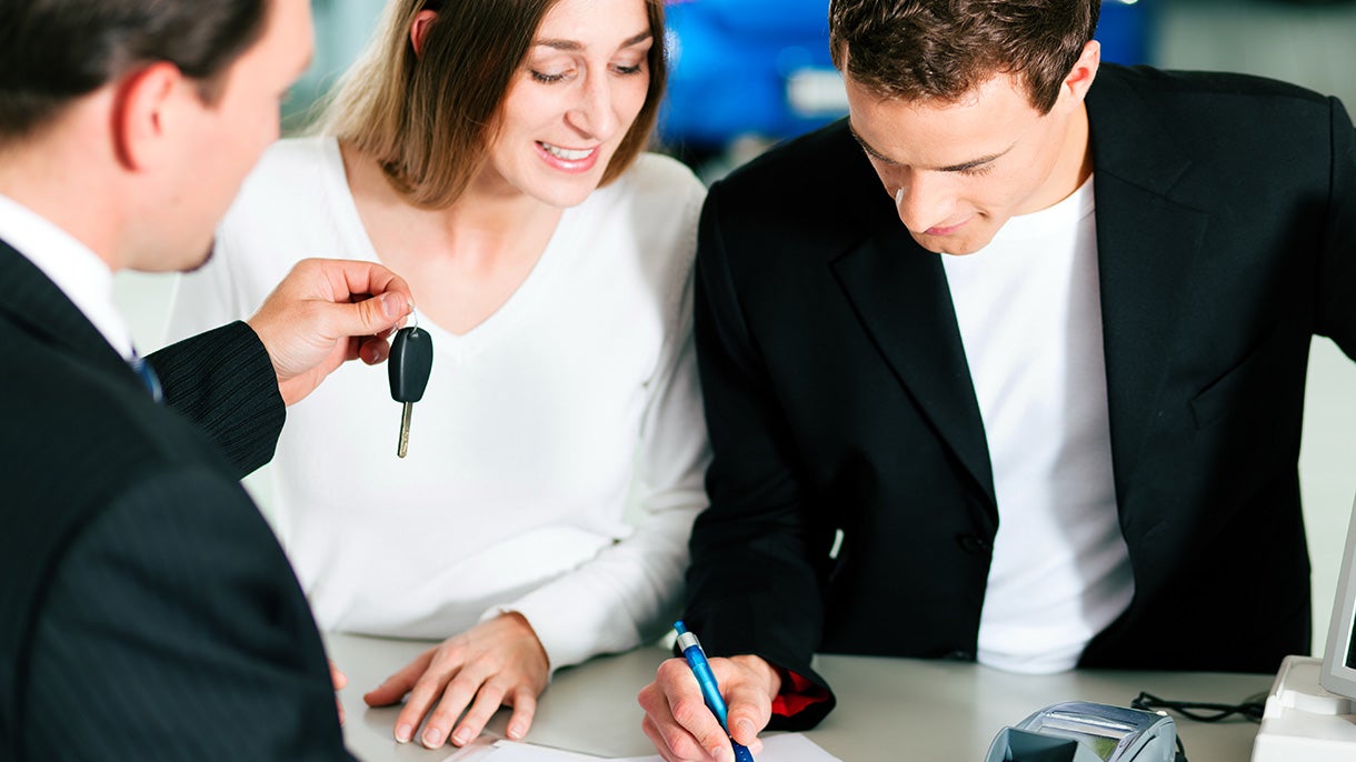 A salesperson in a suit handing car keys to a smiling woman as a man signs paperwork with a blue pen.