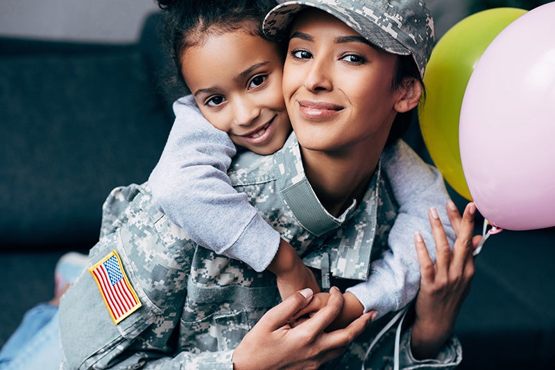 A smiling Black female soldier in uniform holds her smiling Black daughter, who hugs her from behind, with balloons.