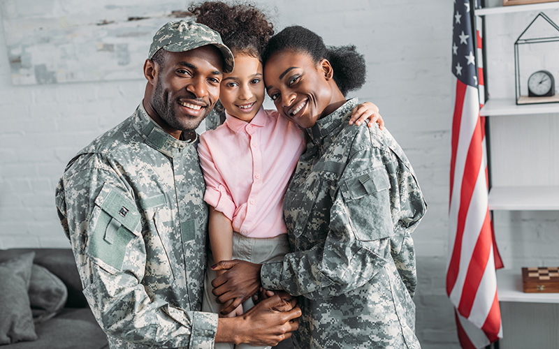A smiling Black military couple in uniform embraces their happy young daughter, with an American flag in the background.