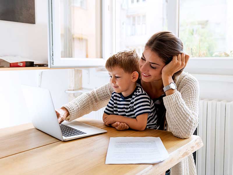 A smiling mother and her young son watch something on a laptop together, resting on a wooden table.